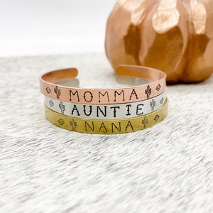 Puede incluir: Tres pulseras apiladas con las palabras "Momma", "Auntie" y "Nana" grabadas en ellas. Cada pulsera tiene un peque&ntilde;o dise&ntilde;o de cactus a cada lado de la palabra.