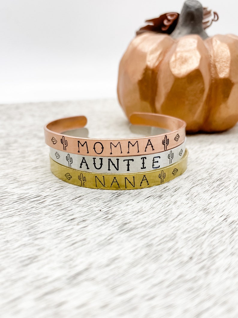 Puede incluir: Tres pulseras apiladas con las palabras "Momma", "Auntie" y "Nana" grabadas en ellas. Cada pulsera tiene un peque&ntilde;o dise&ntilde;o de cactus a cada lado de la palabra.