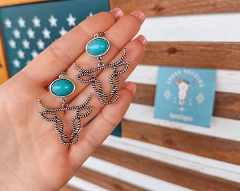 Könnte beinhalten: Ein Paar silberne Ohrringe mit türkisfarbenen Steinen und einem Stierkopfdesign. Die Ohrringe befinden sich auf einer Hand vor einem Holzgrund mit einer amerikanischen Flagge.