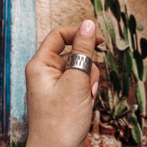 May include: A silver thumb ring with the words "Mama Tried" engraved on it. The ring is worn on a person's thumb.