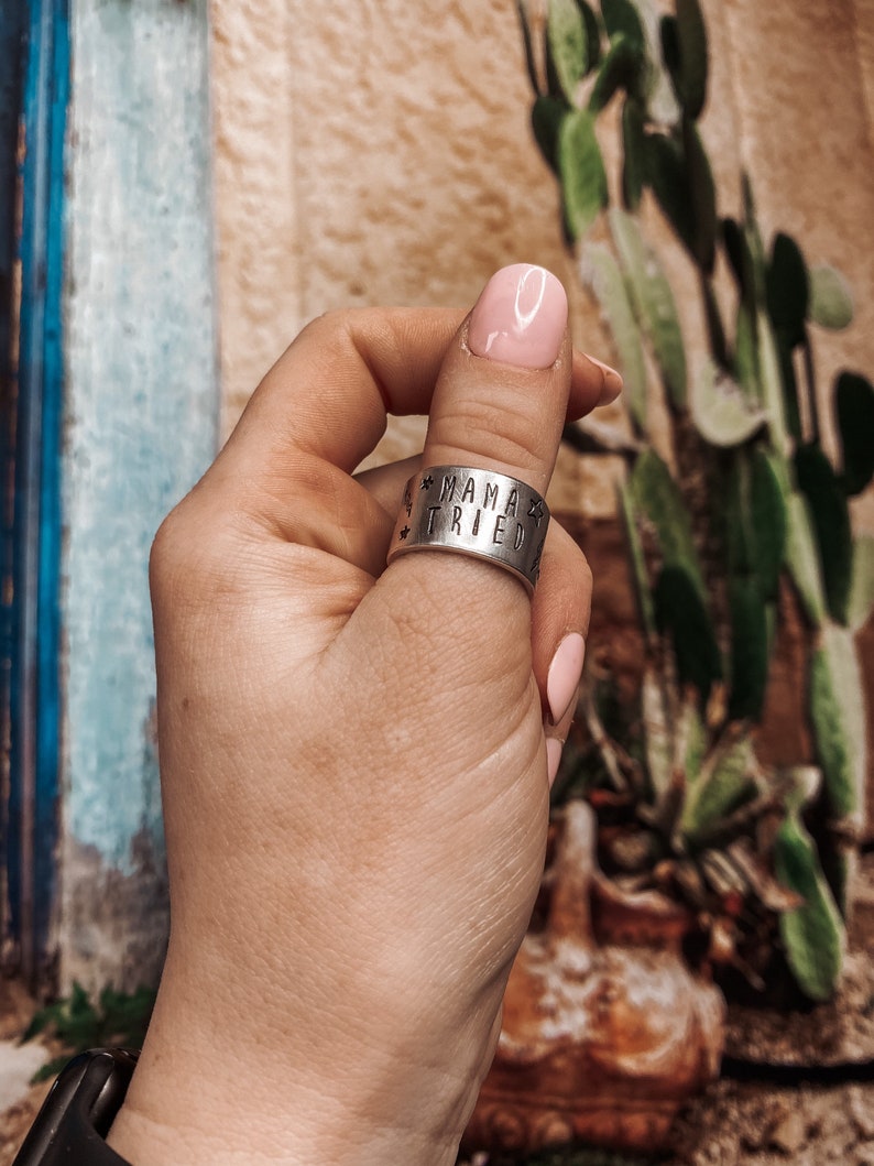 May include: A silver thumb ring with the words "Mama Tried" engraved on it. The ring is worn on a person's thumb.