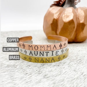 Puede incluir: Tres pulseras apiladas con las palabras "Momma", "Auntie" y "Nana" grabadas en ellas. Cada pulsera es de un metal diferente: cobre, aluminio y lat&oacute;n. Cada pulsera tiene un peque&ntilde;o dise&ntilde;o de cactus a cada lado del texto.