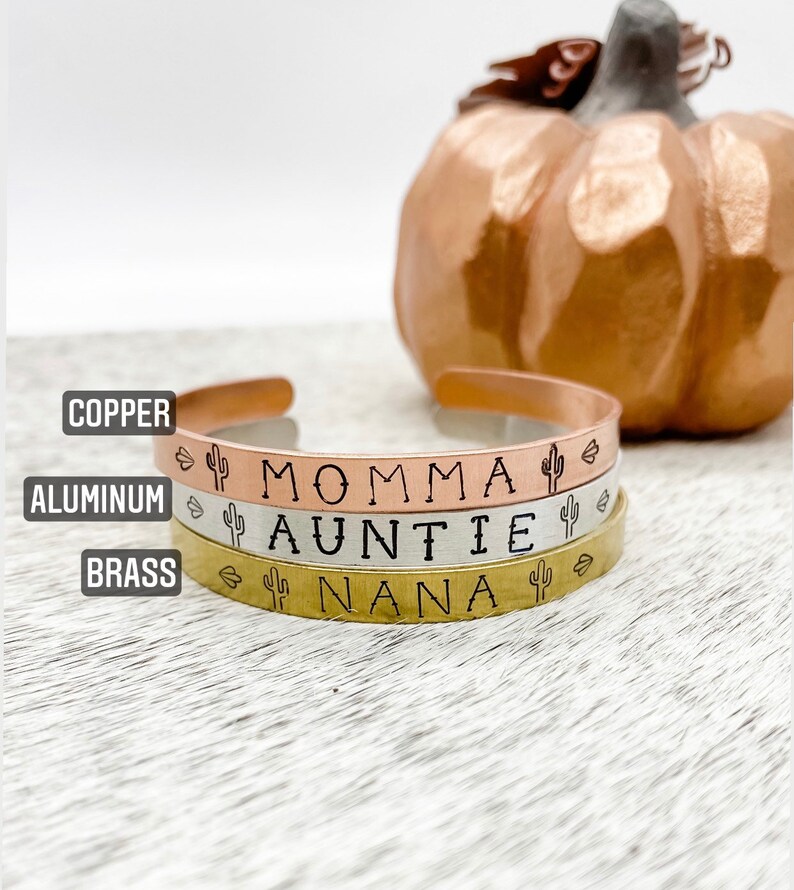 Puede incluir: Tres pulseras apiladas con las palabras "Momma", "Auntie" y "Nana" grabadas en ellas. Cada pulsera es de un metal diferente: cobre, aluminio y lat&oacute;n. Cada pulsera tiene un peque&ntilde;o dise&ntilde;o de cactus a cada lado del texto.
