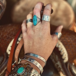 May include: A hand wearing a silver ring with a turquoise stone, a silver band ring engraved with the word "BOSS", and several silver bracelets with engraved designs. The hand is resting on a brown leather surface.