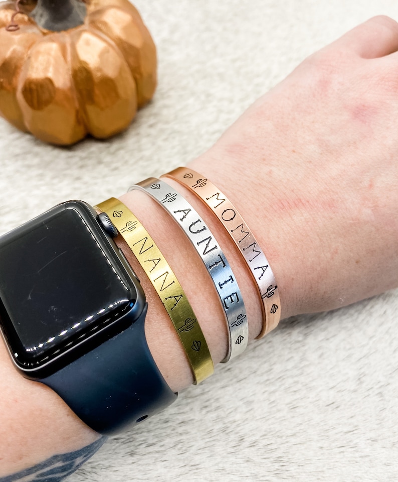Puede incluir: Tres pulseras de oro, plata y oro rosa con un dise&ntilde;o de cactus y coraz&oacute;n. Las pulseras est&aacute;n grabadas con las palabras "MOMMA", "AUNTIE" y "NANA".