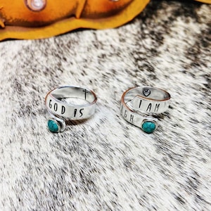 May include: Two silver rings with turquoise stones. The first ring says "GOD IS" with a cross symbol. The second ring says "I AM ENOUGH" with a heart symbol.
