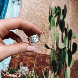 May include: A silver ring with the words "Mama Tribe" engraved on it. The ring is being held by a hand with pink nail polish.