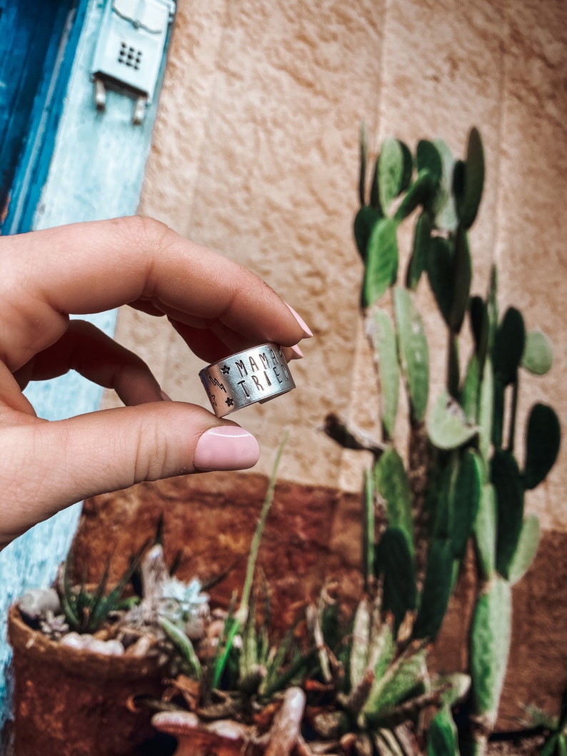 May include: A silver ring with the words "Mama Tribe" engraved on it. The ring is being held by a hand with pink nail polish.