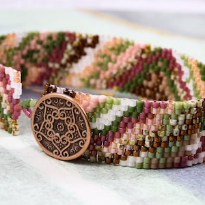 Pode incluir: Um bracelete de contas com um padrão geométrico em tons de rosa, verde e marrom. O bracelete tem um fecho de cobre com um design intrincado.