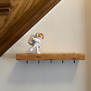 May include: A white astronaut figurine with a gold helmet stands on a wooden shelf with five hooks. The shelf is mounted on a white wall.
