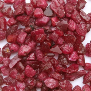 May include: A collection of raw, uncut ruby gemstones. The rubies are a deep, rich red color, with varying shapes and sizes. The gemstones are scattered across a white surface.
