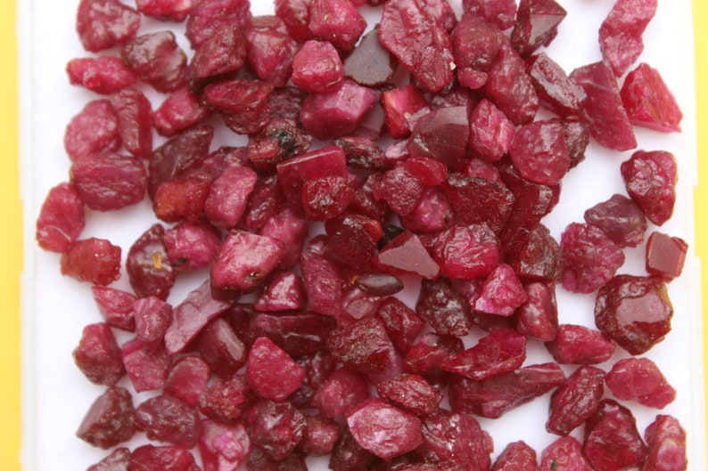 May include: A collection of raw, uncut ruby gemstones. The rubies are a deep, rich red color, with varying shapes and sizes. The gemstones are scattered across a white surface.