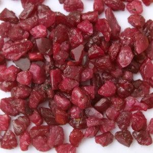 May include: A collection of raw, uncut ruby gemstones. The rubies are a deep, rich red color, with varying shapes and sizes. They are scattered across a white surface, with a yellow background.