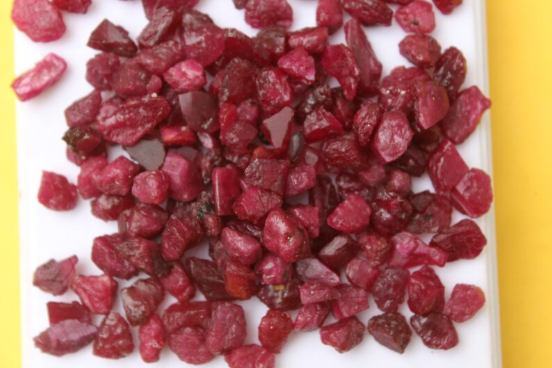 May include: A collection of raw, uncut ruby gemstones. The rubies are a deep, rich red color, with varying shapes and sizes. They are scattered across a white surface, with a yellow background.