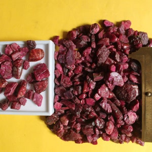 Puede incluir: Una colección de piedras preciosas de rubí en bruto, sin cortar, de varios tamaños y formas, expuestas en una bandeja blanca y un montón. Una regla con texto se coloca entre los rubíes. El fondo es amarillo liso.