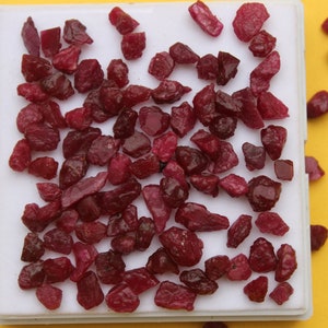 May include: A collection of raw, uncut ruby gemstones in varying shapes and sizes. The deep red stones are displayed on a white tray against a bright yellow background. These natural gemstones are ideal for jewelry making or collecting.