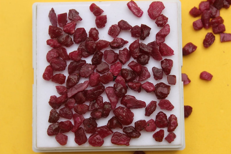 May include: A collection of raw, uncut ruby gemstones in varying shapes and sizes. The deep red stones are displayed on a white tray against a bright yellow background. These natural gemstones are ideal for jewelry making or collecting.