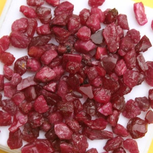 May include: A collection of raw, uncut ruby gemstones. The rubies vary in size and shape, displaying a deep red color with some darker, more opaque pieces. They are scattered on a white surface against a yellow background.