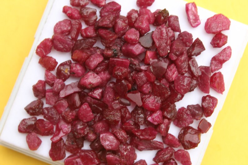 May include: A collection of raw, uncut ruby gemstones. The rubies vary in size and shape, displaying a deep red color with some darker, more opaque pieces. They are scattered on a white surface against a yellow background.