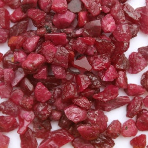 May include: A collection of raw, uncut ruby gemstones. The stones are a deep, rich red color, with irregular shapes and varying sizes. The gemstones are scattered across a white surface.