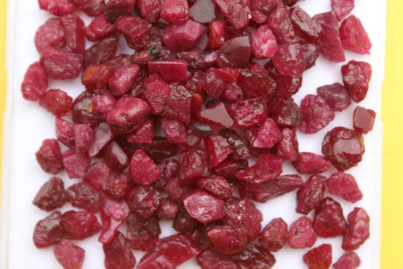 May include: A collection of raw, uncut ruby gemstones. The stones are a deep, rich red color, with irregular shapes and varying sizes. The gemstones are scattered across a white surface.