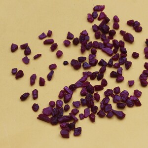 May include: A collection of small, rough-cut ruby gemstones in varying shapes and sizes. The deep red-purple stones are scattered on a yellow surface, with a metal measuring tool in the corner.