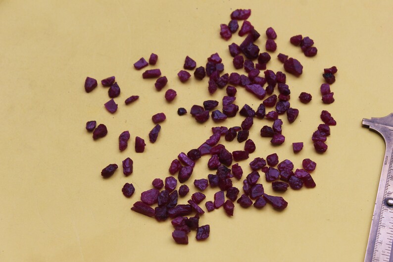 May include: A collection of small, rough-cut ruby gemstones in varying shapes and sizes. The deep red-purple stones are scattered on a yellow surface, with a metal measuring tool in the corner.