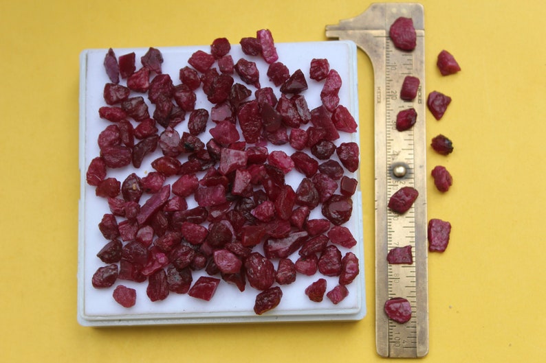 May include: A collection of raw, uncut ruby gemstones in varying sizes and shapes, displayed on a white tray. A brass ruler is positioned next to the gemstones for scale, with a bright yellow background.