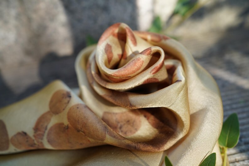Pu&ograve; includere: Una sciarpa in tessuto liscio color oro con un motivo ripetitivo di stampe botaniche arrotondate color ruggine. Il materiale &egrave; piegato in una forma a rosa a strati, che mette in risalto la lucentezza morbida e le macchie organiche color marrone terra.