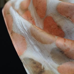 May include: A lightweight scarf featuring a cream-colored base with a repeating pattern of soft, rounded peach and muted brown botanical-style prints. The fabric has a smooth, slightly lustrous texture with visible folds that create depth and dimension.