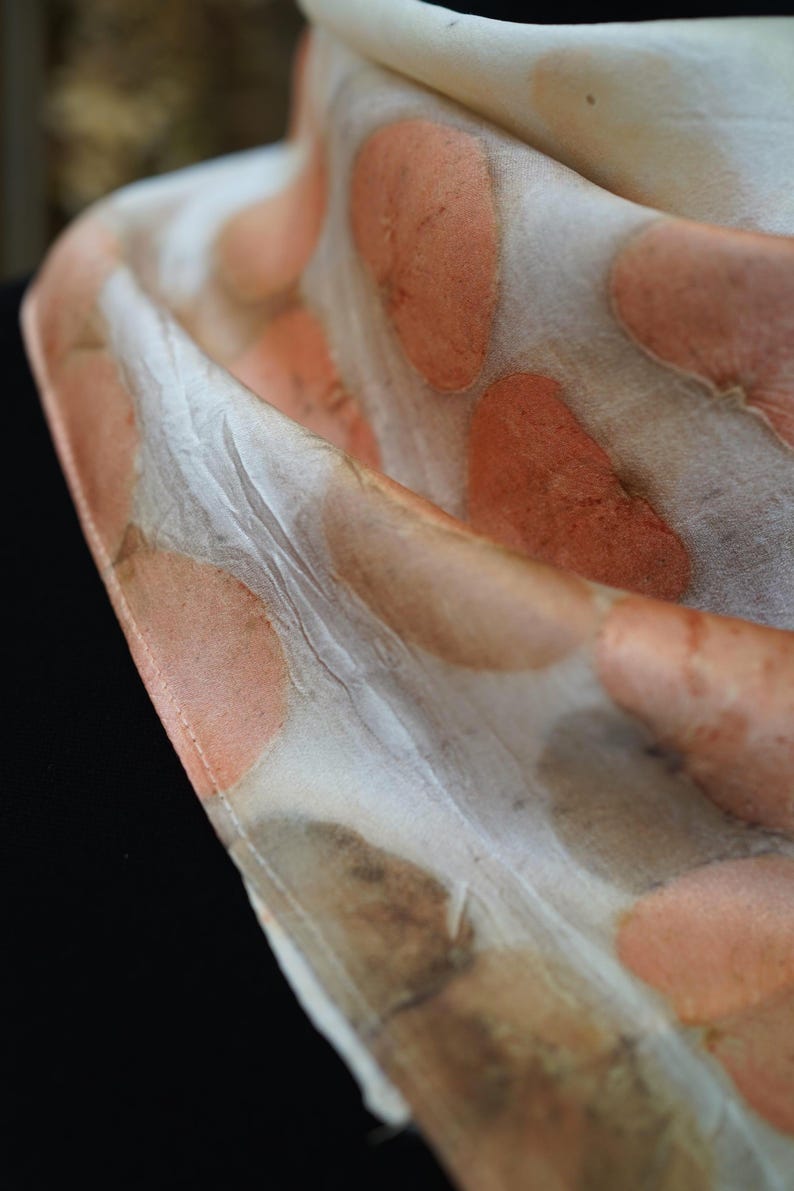 May include: A lightweight scarf featuring a cream-colored base with a repeating pattern of soft, rounded peach and muted brown botanical-style prints. The fabric has a smooth, slightly lustrous texture with visible folds that create depth and dimension.