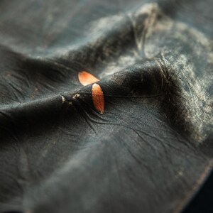 May include: A dark charcoal gray fabric with a wrinkled, textured surface featuring a subtle, faded white pattern and two small, smooth, copper-colored oval accents resting on a fold in the center of the material.