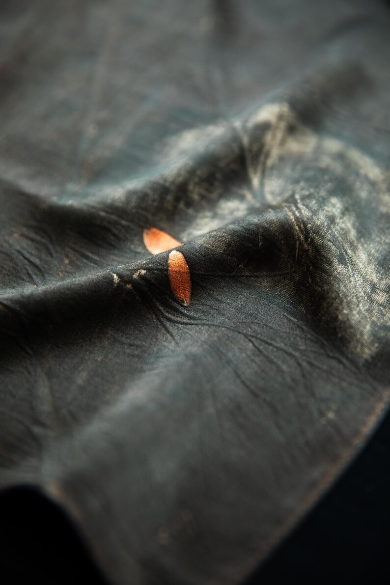 May include: A dark charcoal gray fabric with a wrinkled, textured surface featuring a subtle, faded white pattern and two small, smooth, copper-colored oval accents resting on a fold in the center of the material.