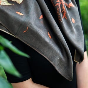 May include: A square scarf in a dark charcoal gray hue featuring botanical prints of orange and rust-colored leaves and stems. The fabric displays a subtle texture and is draped around the neck, showcasing a natural leaf pattern across the surface.