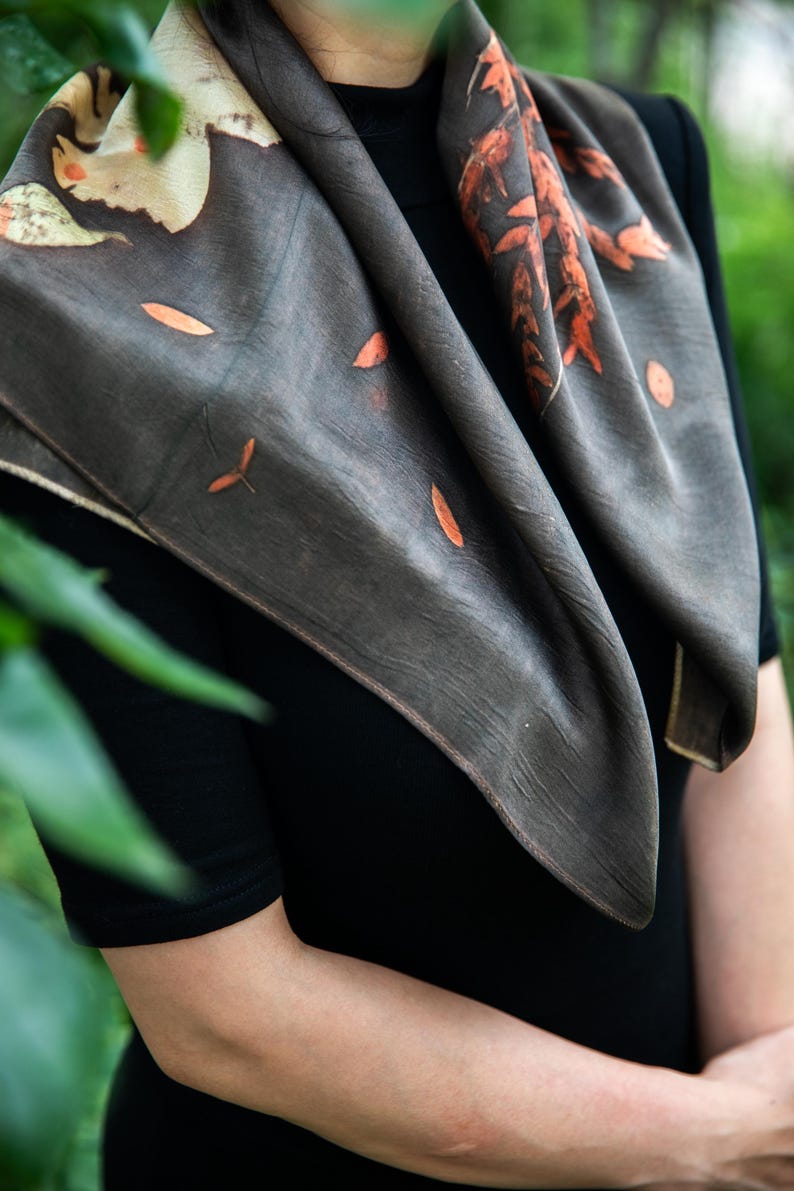 May include: A square scarf in a dark charcoal gray hue featuring botanical prints of orange and rust-colored leaves and stems. The fabric displays a subtle texture and is draped around the neck, showcasing a natural leaf pattern across the surface.