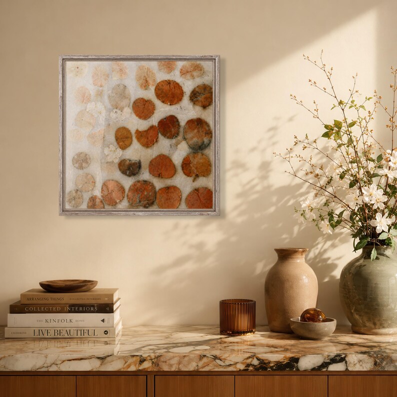May include: A square framed wall art piece featuring a white background with a scattered pattern of circular, pressed orange and brown dried leaves. The artwork is enclosed in a thin, light-coloured wooden frame with a textured finish.