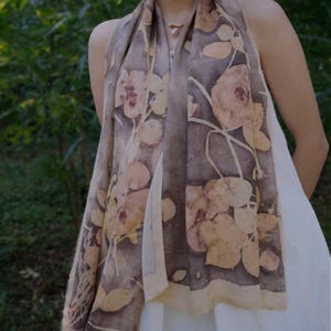 May include: A long, rectangular scarf with a floral pattern in shades of brown, beige, and cream. The scarf is draped over the shoulders, showcasing the botanical print. The background is a blurred green, suggesting an outdoor setting.