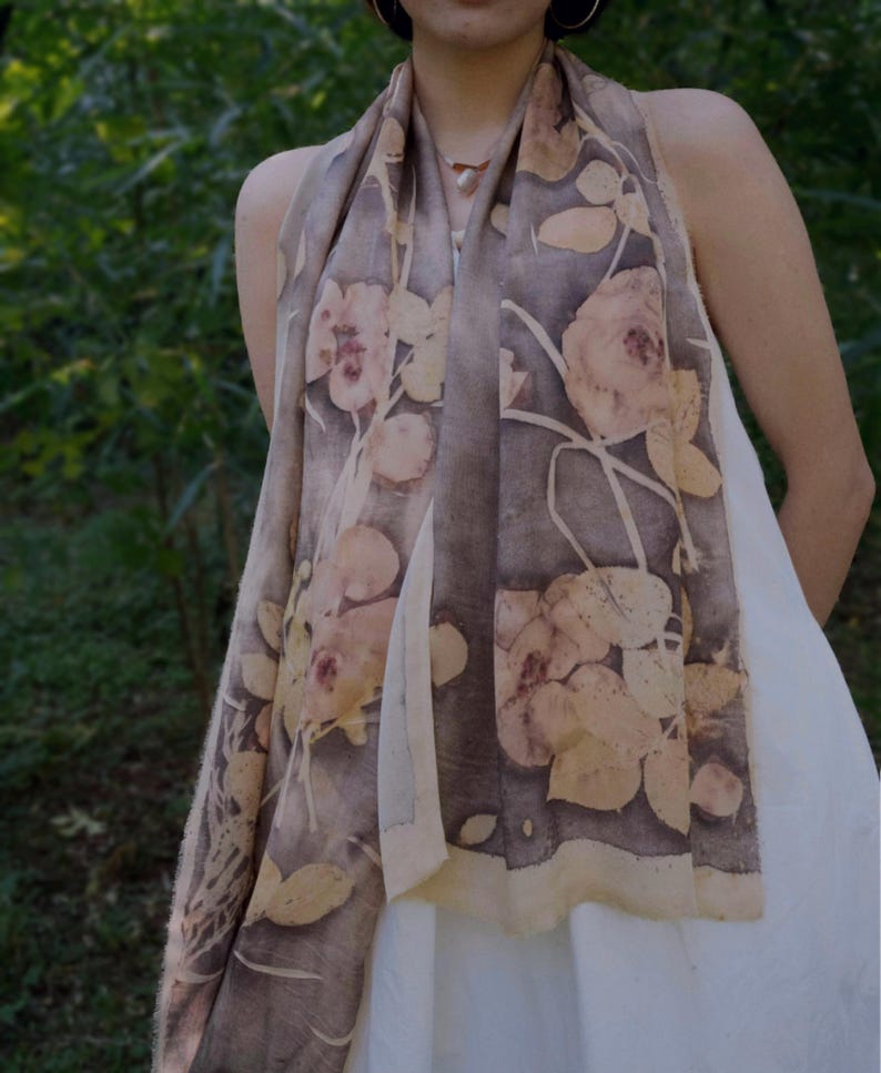 May include: A long, rectangular scarf with a floral pattern in shades of brown, beige, and cream. The scarf is draped over the shoulders, showcasing the botanical print. The background is a blurred green, suggesting an outdoor setting.