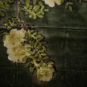 May include: A dark olive green fabric featuring a botanical print of white flowers and green leaves. The design displays a branch with detailed foliage and blossoms, with dark outlines surrounding the plant elements against the solid background.