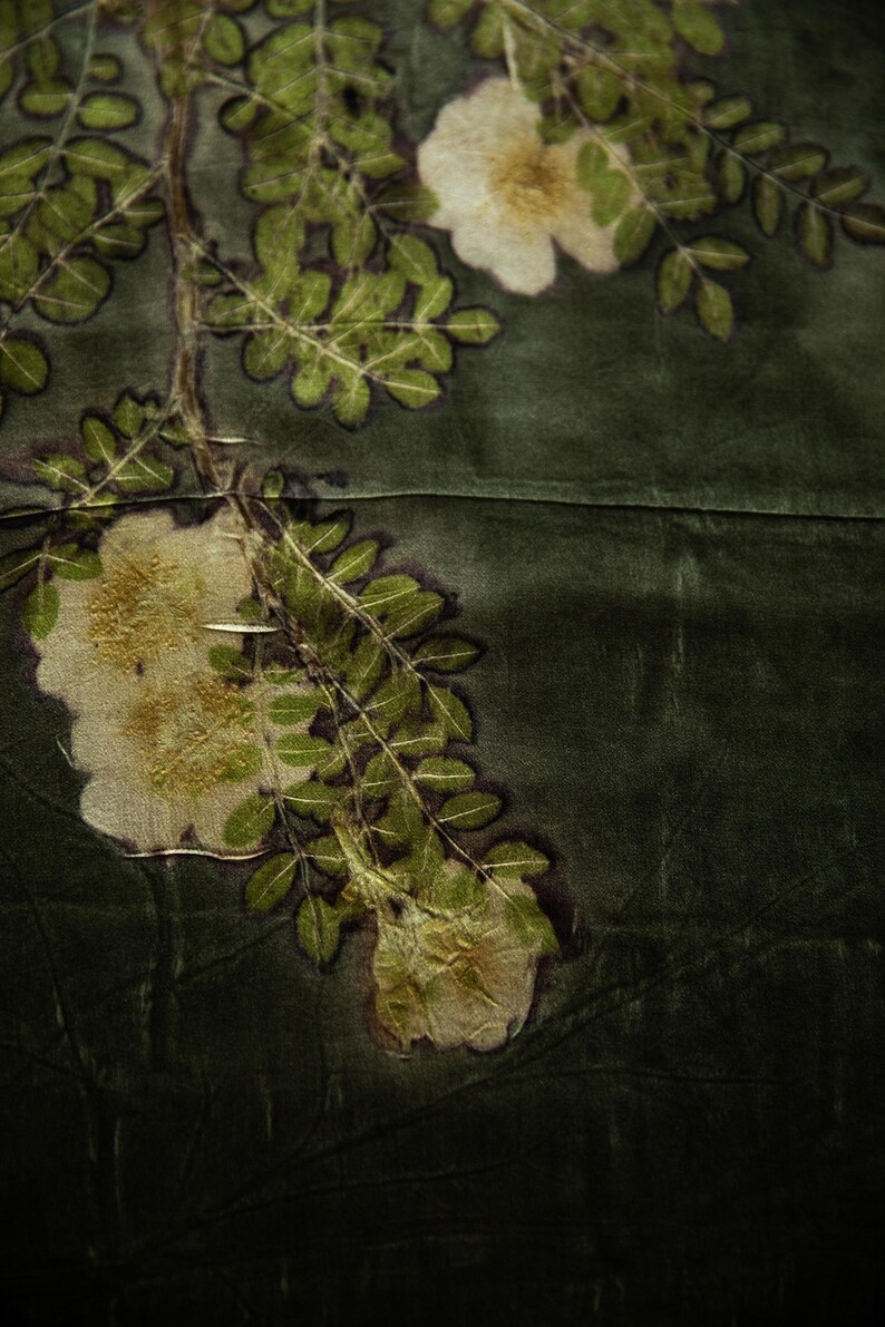 May include: A dark olive green fabric featuring a botanical print of white flowers and green leaves. The design displays a branch with detailed foliage and blossoms, with dark outlines surrounding the plant elements against the solid background.