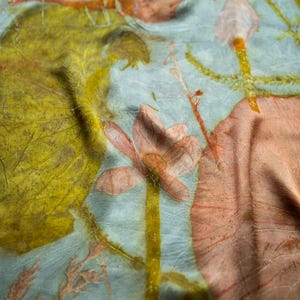 May include: A light blue fabric featuring botanical prints in shades of mustard yellow and muted orange. The textile displays detailed imprints of large leaves, slender stems, and a central flower, creating a layered, organic pattern across the surface.