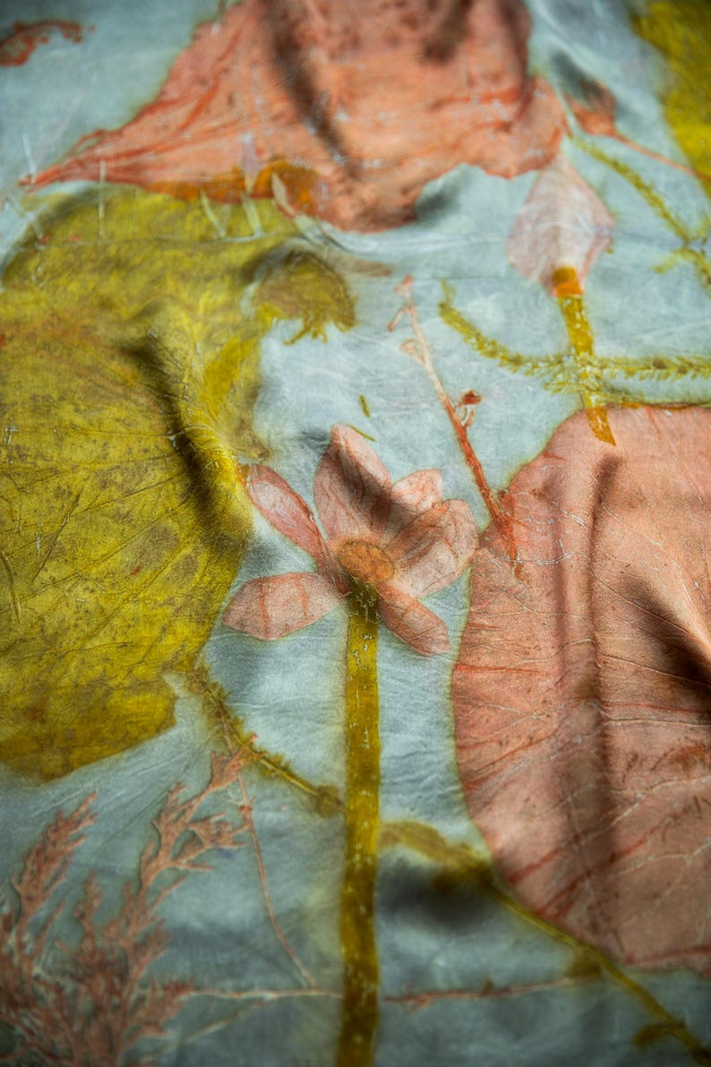 May include: A light blue fabric featuring botanical prints in shades of mustard yellow and muted orange. The textile displays detailed imprints of large leaves, slender stems, and a central flower, creating a layered, organic pattern across the surface.