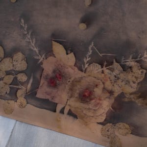 May include: A textile piece featuring a botanical print with dark grey and charcoal tones. The fabric displays light beige leaf and floral imprints, including rose-like shapes with small, distinct red centres, arranged in a natural, organic pattern.