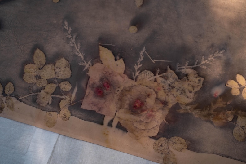May include: A textile piece featuring a botanical print with dark grey and charcoal tones. The fabric displays light beige leaf and floral imprints, including rose-like shapes with small, distinct red centres, arranged in a natural, organic pattern.
