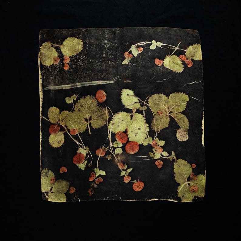 May include: A square textile piece featuring a dark black background with a botanical print of green strawberry leaves and reddish-brown berries scattered across the surface. The design displays natural plant imprints with visible veins.