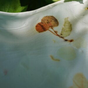 May include: A white fabric featuring botanical imprints of leaves in shades of orange, brown, and pale green. The material has a soft, draped texture with visible folds and creases, showcasing natural plant-based pigment patterns on the surface.