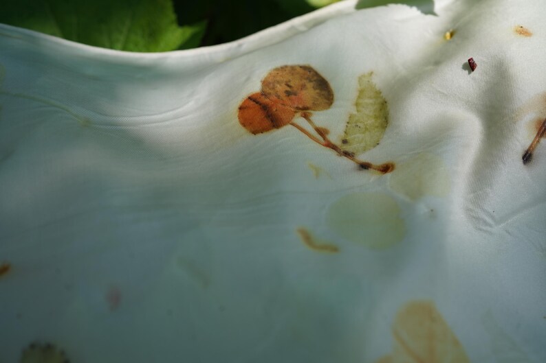 May include: A white fabric featuring botanical imprints of leaves in shades of orange, brown, and pale green. The material has a soft, draped texture with visible folds and creases, showcasing natural plant-based pigment patterns on the surface.