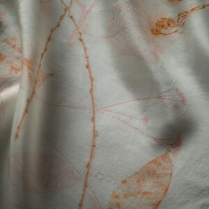 May include: A white fabric scarf featuring a botanical print with delicate orange and soft pink leaf and stem patterns. The textile has a smooth, slightly lustrous surface with natural folds that create depth and highlight the organic design.
