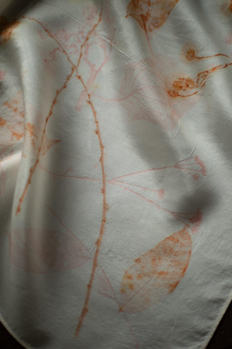 May include: A white fabric scarf featuring a botanical print with delicate orange and soft pink leaf and stem patterns. The textile has a smooth, slightly lustrous surface with natural folds that create depth and highlight the organic design.