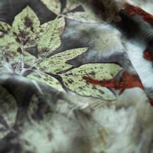 May include: A lightweight fabric featuring a botanical print with large, detailed green leaves and dark brown, mottled background tones. The textile displays organic, earthy patterns with subtle accents of deep red throughout the surface.